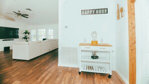 A modern living room with a white sofa, wooden flooring, a ceiling fan, a wall-mounted TV, and a 'HAPPY HOUR' sign.
