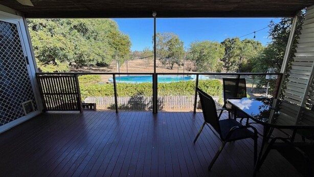 A covered outdoor deck with a view of a swimming pool, surrounded by trees and a fence.