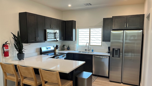 Una cocina moderna con gabinetes oscuros, electrodomésticos de acero inoxidable, una isla central con sillas y un ventanal que proporciona luz natural.