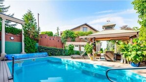 Ein Schwimmbad mit Holzterrasse, einer überdachten Sitzgelegenheit und einem Haus im Hintergrund.