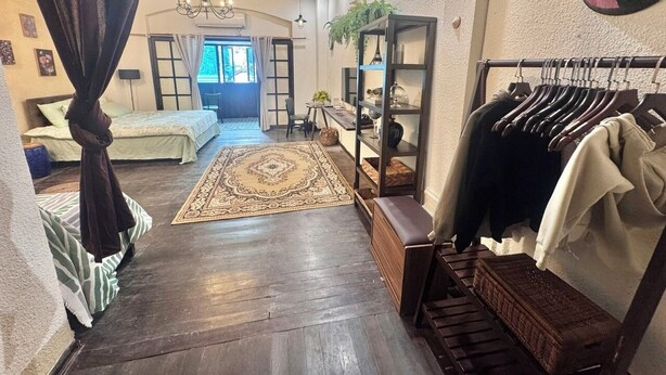A spacious living area with a dark wooden floor, a large rug, a sofa, a coffee table, a bookshelf, and a clothing rack.