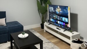 A living room with a flat-screen TV, a clock, a sofa, a coffee table, and a plant. - Charming 1-bedroom apartment in enchanting Moncton with WiFi (Moncton)