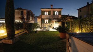 A two-story house with a stone facade, green shutters, and a well-lit front yard with a palm tree and potted plants. - Casa I Tre Cipressi with Swimming Pool (Capannori)