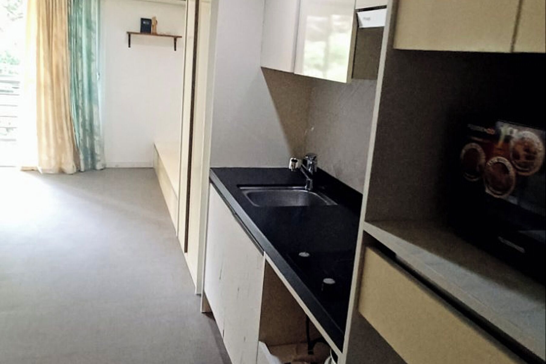 Cucina con piano di lavoro nero, lavello e pensili. Si nota uno scaffale con un libro e una piccola pianta. Il pavimento è chiaro e c'è una tenda alla finestra.