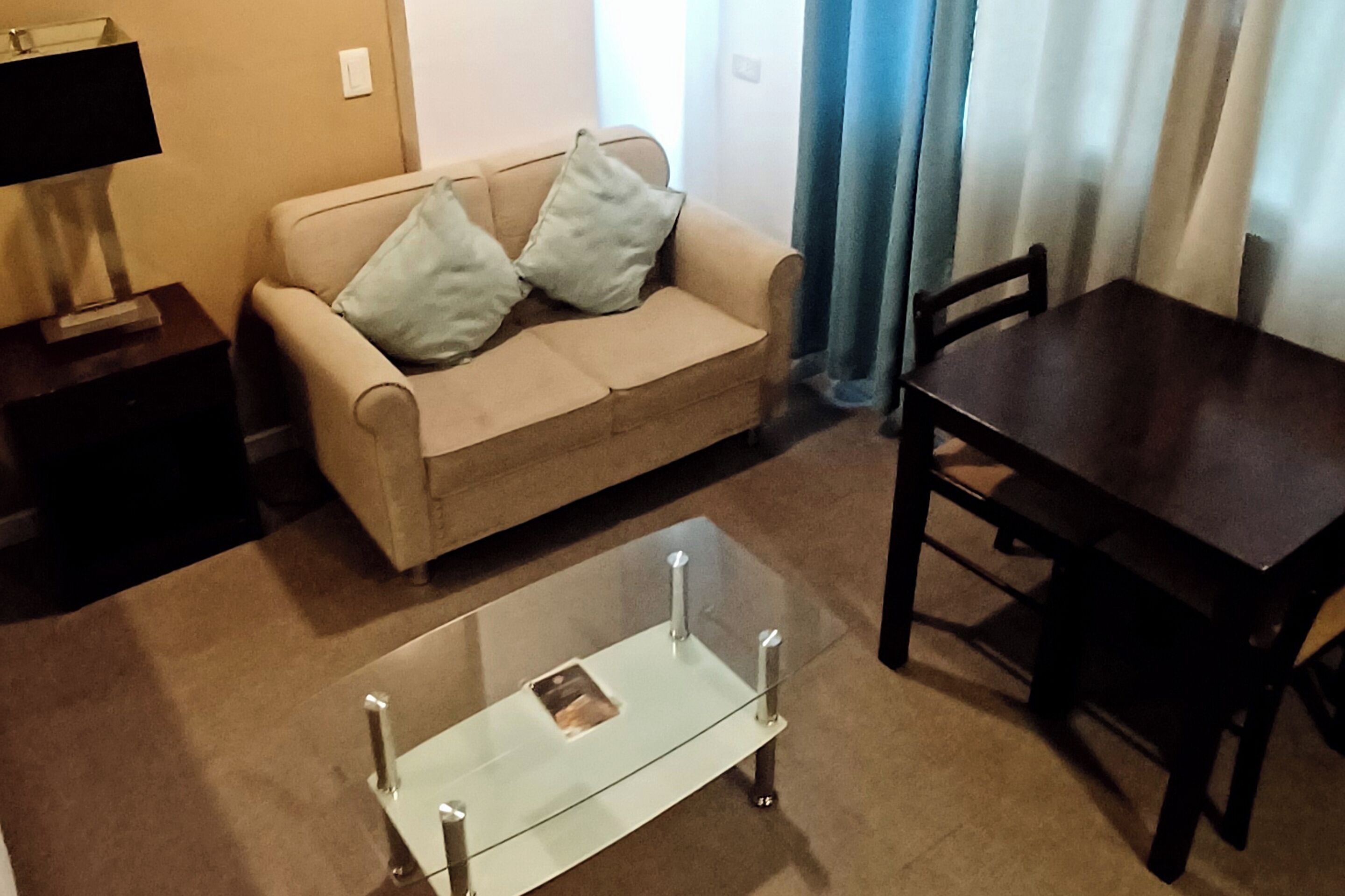 Una sala de estar con un sofá beige, dos cojines, una mesa de centro de vidrio, una mesa de comedor de madera oscura y una lámpara sobre una mesita auxiliar.