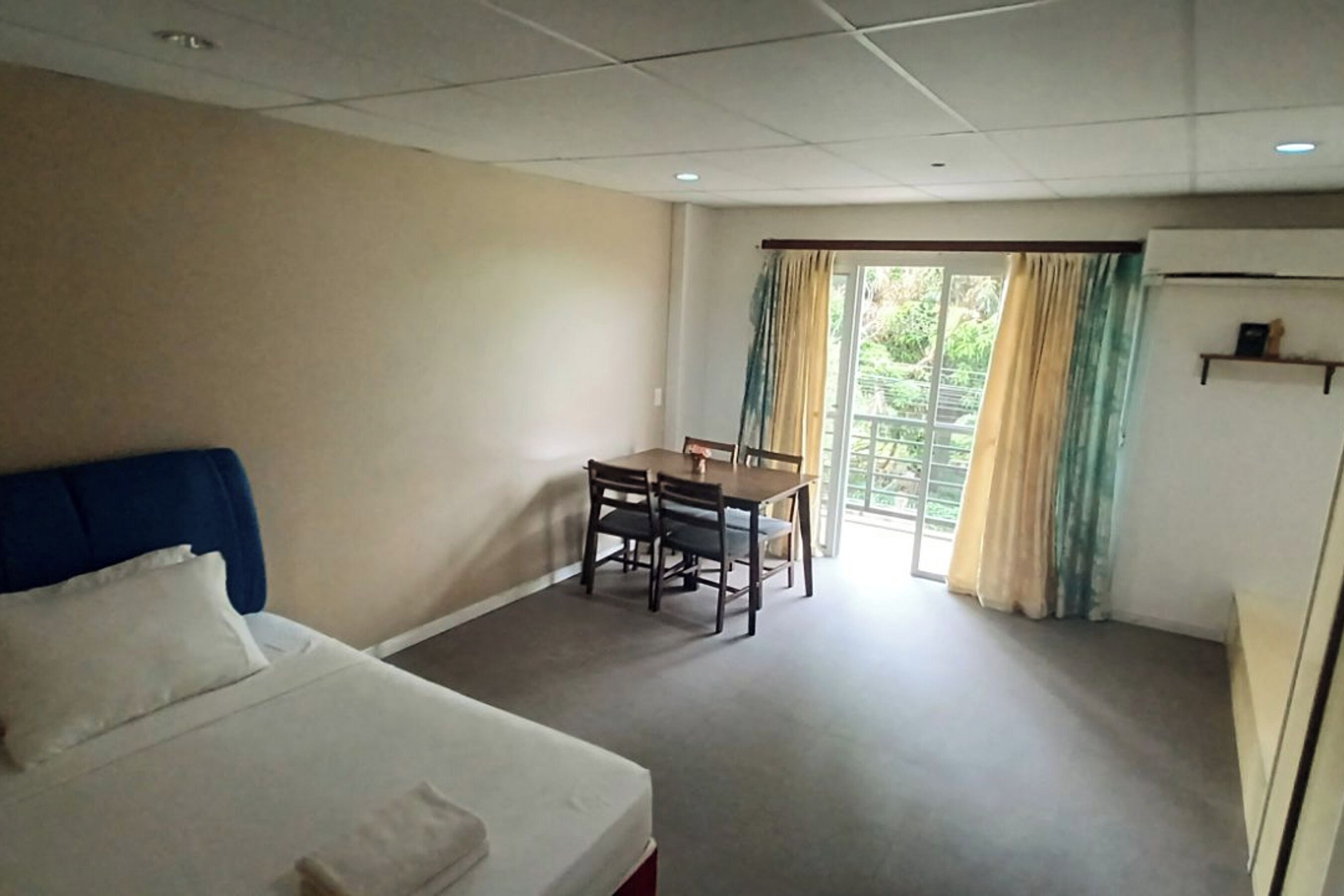 A hotel room with a bed, a dining table with chairs, a small table with a vase, and a window with curtains.