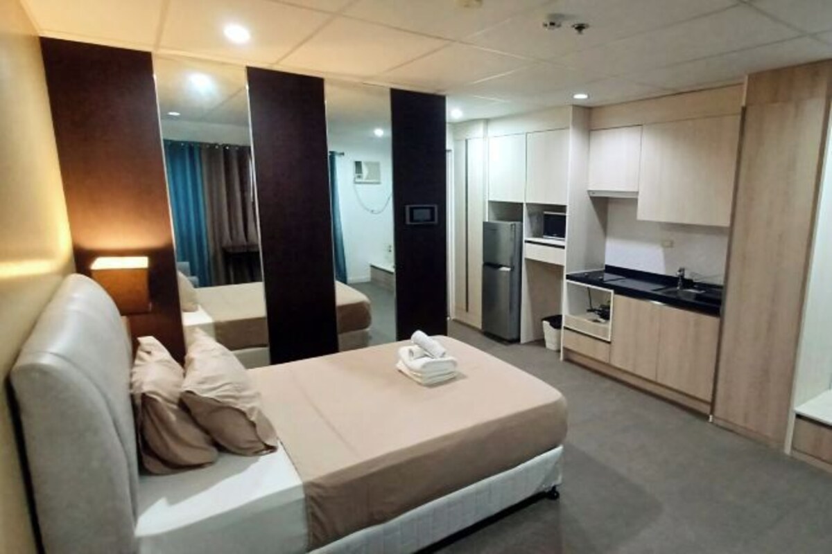 A hotel room with a bed, a small kitchenette, and a bathroom visible in the background.