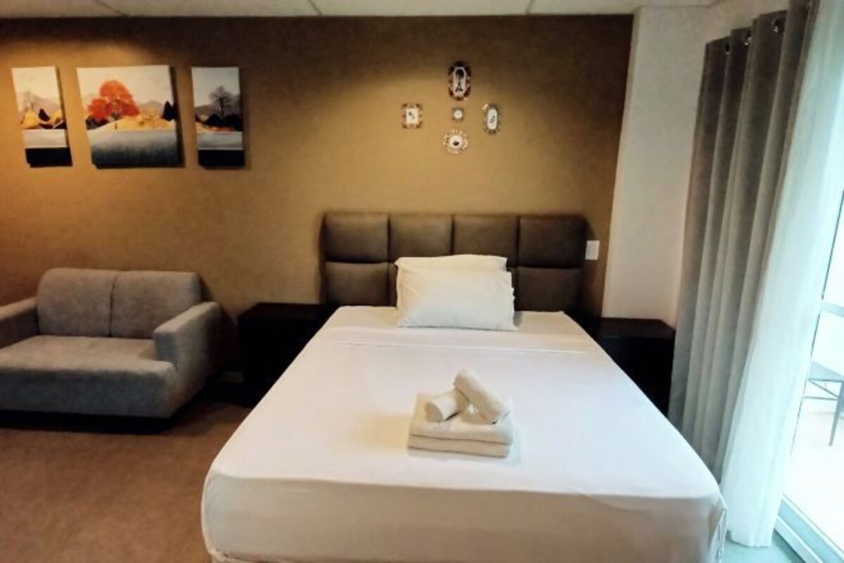 A hotel room with a bed, a grey sofa, and three wall-mounted paintings.