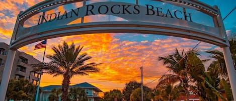 A sign reading 'Indian Rocks Beach' with palm trees and a sunset in the background.