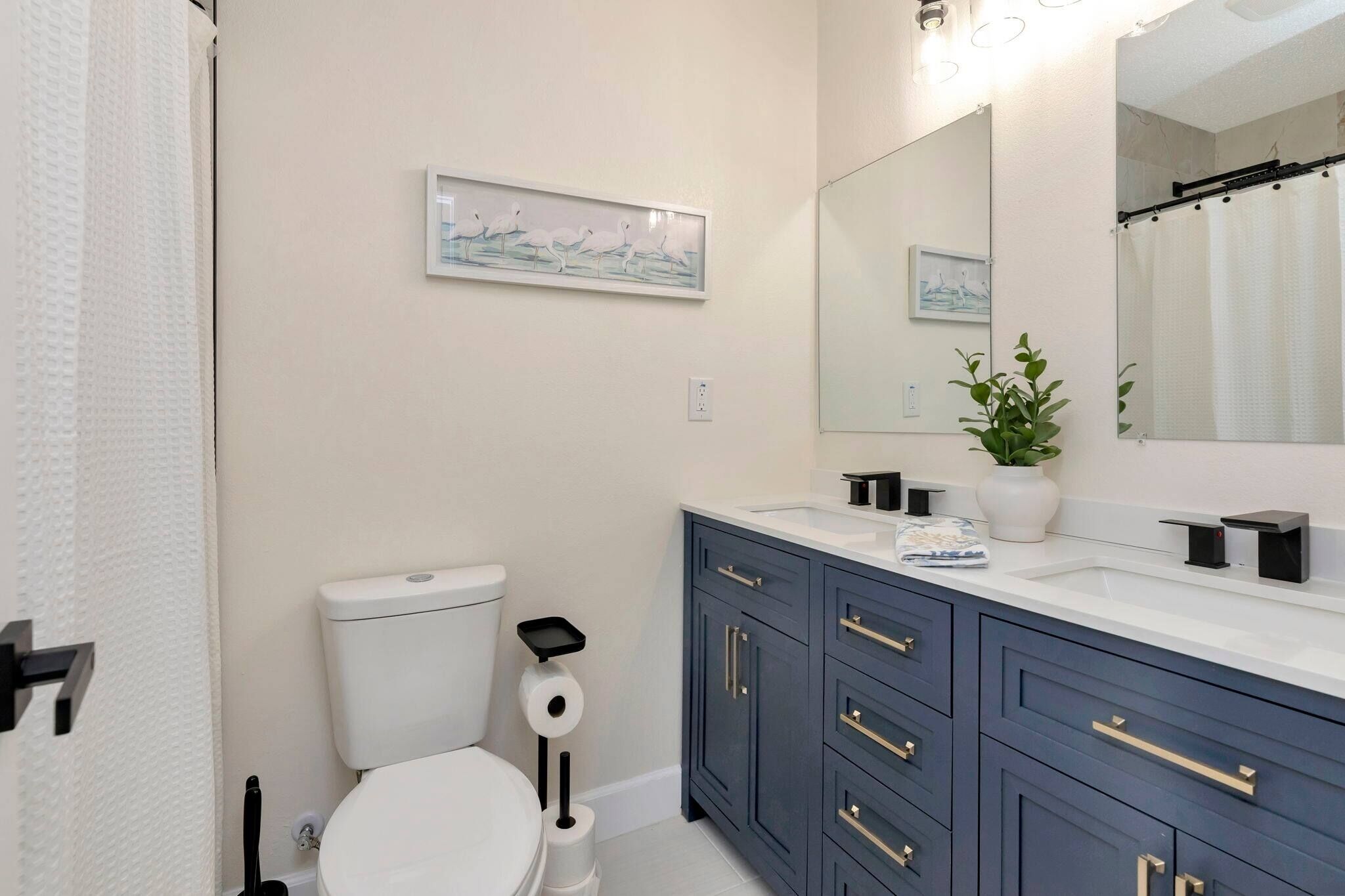 Une salle de bain comprenant des toilettes, un lavabo, un miroir, un tableau encadré accroché au mur et une plante posée sur le comptoir.