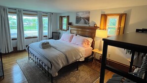 Ein Schlafzimmer mit Bett, Nachttisch, Lampe, Heizkörper und Blick ins Freie durch die Fenster.