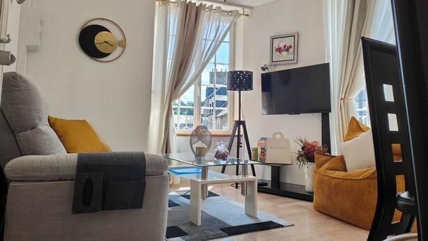 A modern living room with a sofa, a glass coffee table, a TV, and a view of buildings outside.