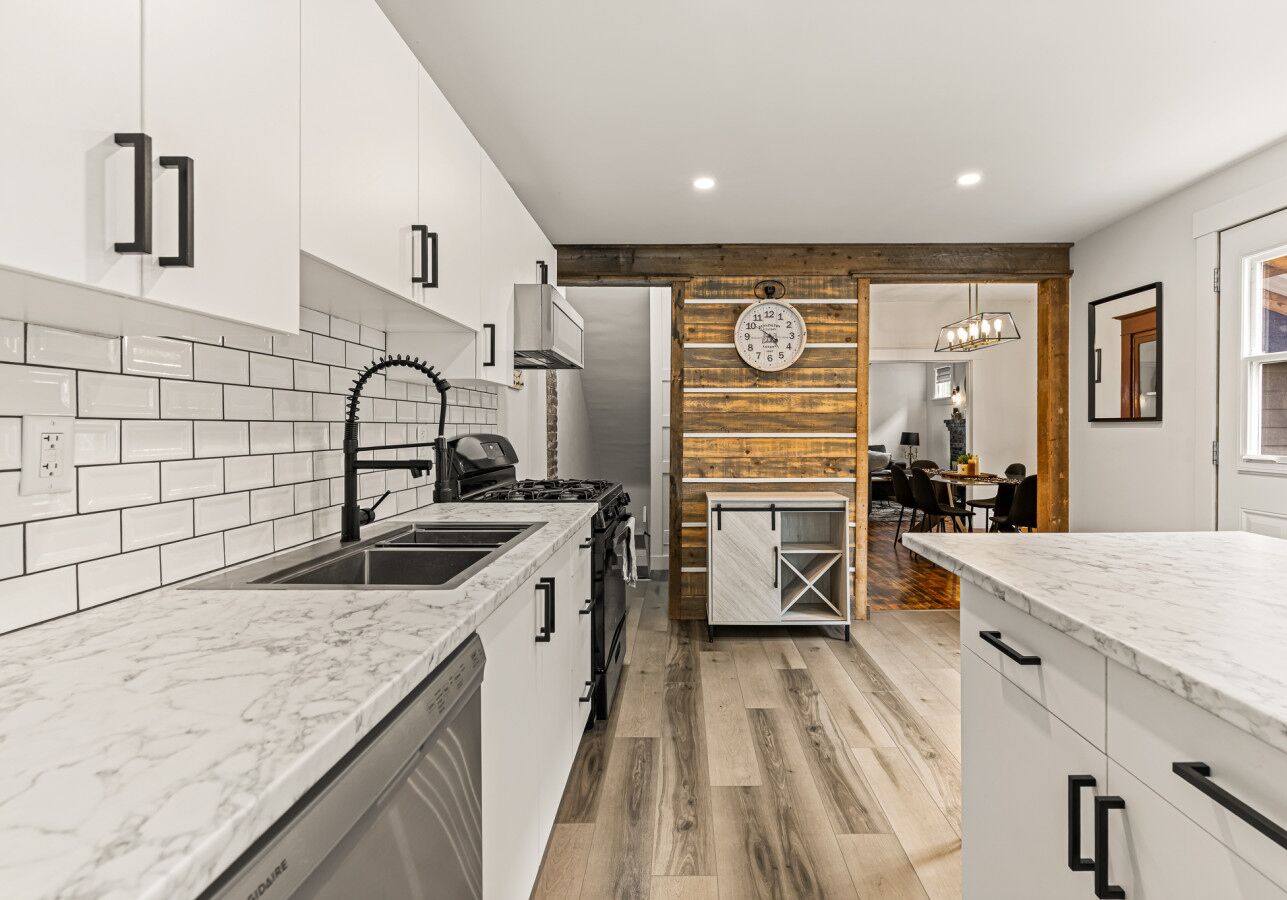 A modern kitchen with white cabinets, a marble countertop, a wooden accent wall, and a dining area in the background.