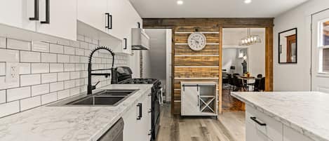 A modern kitchen with white cabinets, a marble countertop, a wooden accent wall, and a dining area in the background.
