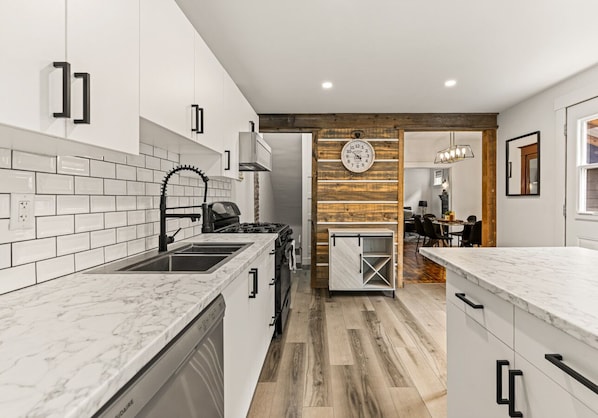 Una cocina moderna con armarios blancos, encimera de mármol, pared decorativa de madera y una zona de comedor al fondo.