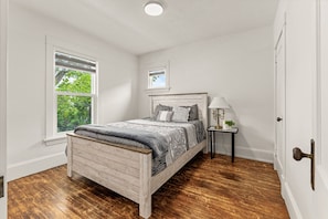 Een slaapkamer met een houten bed, een nachtkastje met een lamp, een raam met jaloezieën en een deur.