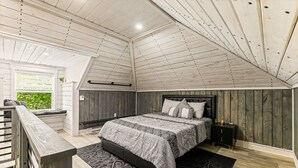 Une chambre avec un plafond en pente, un lit recouvert d’une couvre-lit à motifs, une table de chevet et une fenêtre donnant sur un paysage verdoyant.