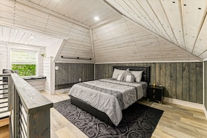 A bedroom with a sloped ceiling, a bed with a patterned bedspread, a nightstand, and a window with a view of greenery. - The Big City Stay | Sleeps 11 | Walkable Location (Moncton)