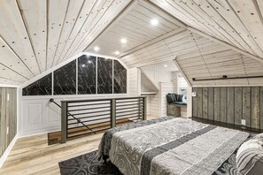 Une chambre dotée d’une grande fenêtre, de poutres apparentes en bois au plafond, d’un lit avec une literie à motifs et d’un petit coin salon avec un banc.