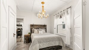Un dormitorio con cabecera de madera, colcha estampada, candelabro y ventana con cortinas traslúcidas.