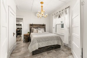 A bedroom with a wooden headboard, a patterned bedspread, a chandelier, and a window with sheer curtains.