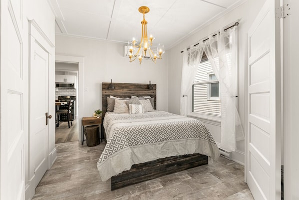 Une chambre à coucher avec une tête de lit en bois, une couvre-lit à motifs, un lustre et une fenêtre ornée de rideaux légers.