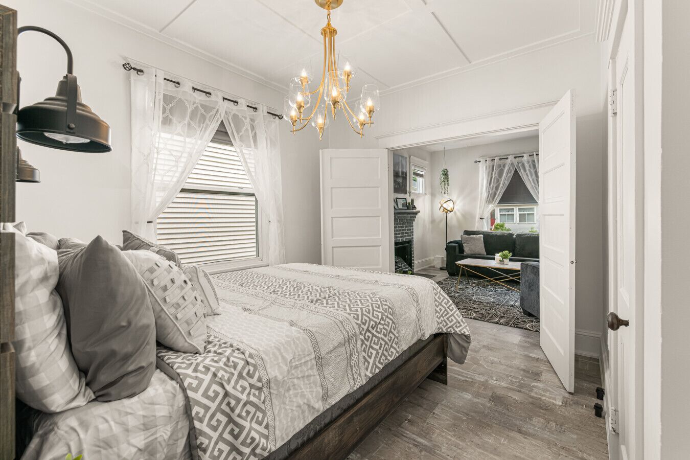 Un dormitorio con una cama grande, una araña de cristal, una ventana con cortinas translúcidas y vistas a otra habitación.