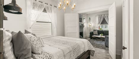 A bedroom with a large bed, a chandelier, a window with sheer curtains, and a view into another room.