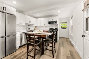Une cuisine moderne avec des meubles blancs, des appareils en acier inoxydable, une table à manger en bois et une vue sur la verdure à l’extérieur.