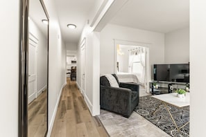 Un salon moderne avec un canapé, une table basse et un téléviseur. Un couloir mène à la cuisine et à la chambre.