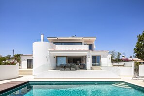 Una casa moderna de dos pisos con piscina, mobiliario exterior y un cielo azul despejado.