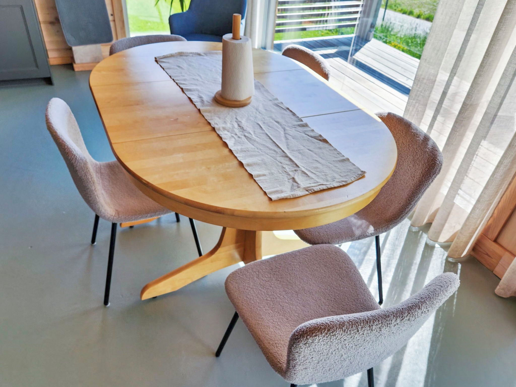 A round wooden dining table with a beige placemat, surrounded by four chairs with a textured fabric seat.