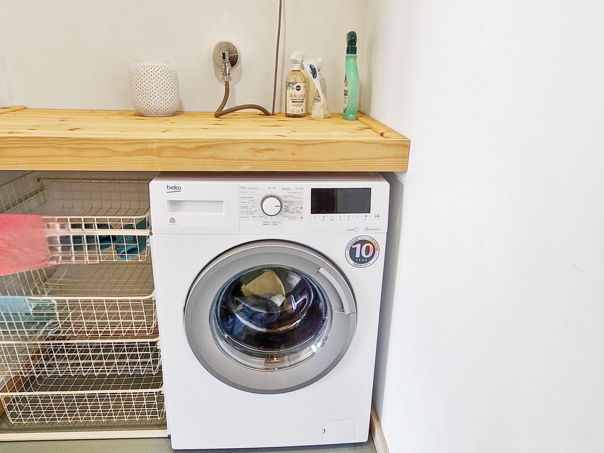 A front-loading washing machine with a control panel, a wooden shelf above it, and a metal rack to the left.