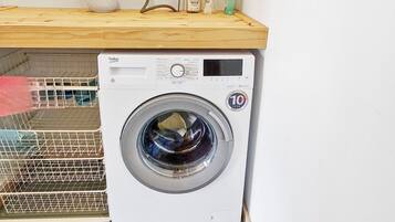 A front-loading washing machine with a control panel, a wooden shelf above it, and a metal rack to the left.