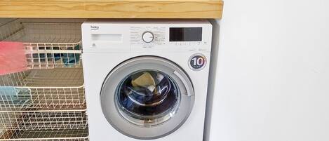 Un lave-linge à chargement frontal doté d’un panneau de commande, d’une étagère en bois au-dessus et d’une grille métallique sur le côté gauche.