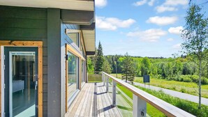 Une terrasse en bois avec des portes vitrées donnant sur une pièce, d’où l’on a vue sur une route et sur la verdure.