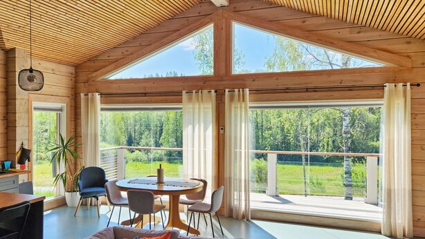 Une pièce spacieuse avec un plafond haut, des poutres en bois, un coin repas avec une table et des chaises, un canapé agrémenté de coussins colorés et de grandes fenêtres offrant une vue sur l’extérieur.