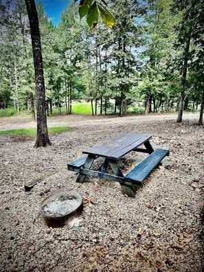 Une table de pique-nique située dans une zone boisée, avec des arbres et un sol recouvert de gravier.