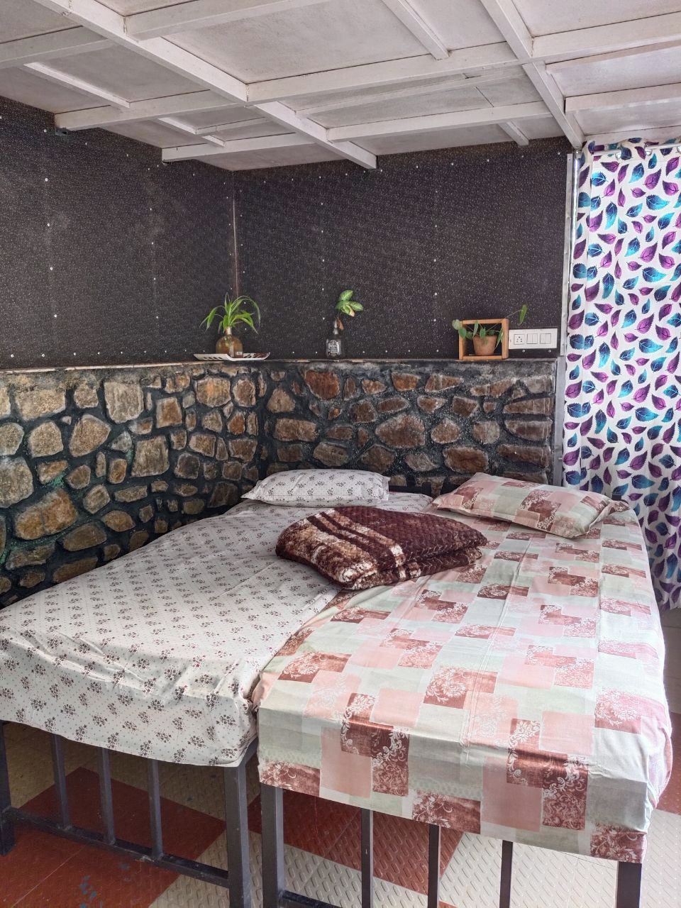 A small, cozy room with a stone wall, a bed with floral bedding, and a window with a leaf-patterned curtain.