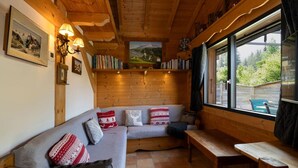 Un interior acogedor de cabaña con techo de madera, un sofá con cojines estampados, un banco de madera y un ventanal con vistas a la vegetación.