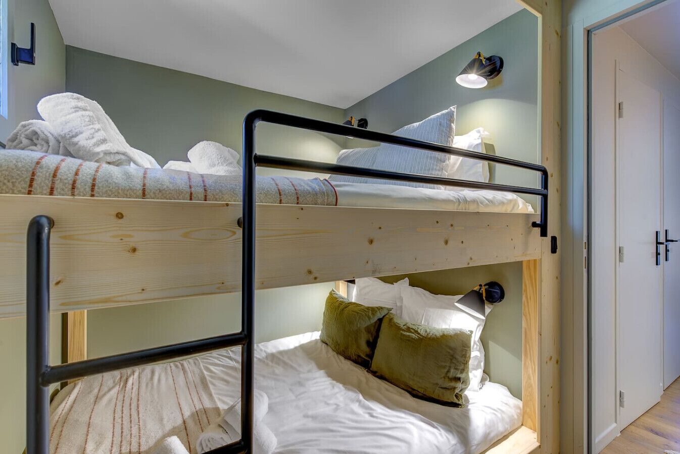 A bunk bed room with two beds, a wooden floor, a white door, and a wall-mounted light fixture.