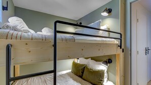 Habitación con literas, dos camas, piso de madera, puerta blanca y una lámpara fijada a la pared.