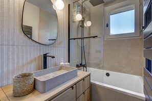Un baño moderno con un lavamanos rectangular, un espejo redondo, una ducha con puerta de cristal y un ventanal que ofrece luz natural.