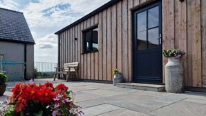 Un edificio in legno con una porta scura e finestre, una piccola zona esterna con una panchina e un tavolo, e una pianta in vaso a terra.