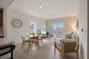 A spacious living area with a dining table, chairs, a sofa, a clock, and large windows with a city view.