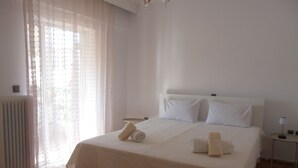 Uma cama bem arrumada com lençóis e almofadas brancas, uma mesinha de cabeceira com luminária e uma janela com cortinas leves.