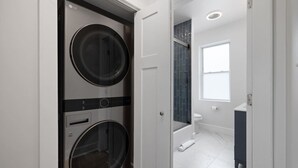 Un cuarto de lavado con dos lavadoras y secadoras de carga frontal, un baño con lavamanos blanco y azulejos oscuros, y una ventana que proporciona luz natural.