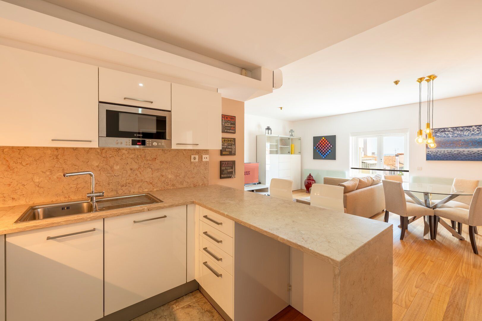 A modern kitchen with white cabinets, a Bosch microwave, and a marble countertop. There is a dining area with a glass table and chairs, and a living space with a sofa and a painting on the wall.