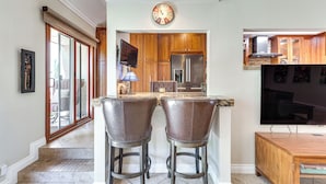 A kitchen with wooden cabinets, a stainless steel refrigerator, and a built-in oven. There is a bar area with two stools and a television mounted on the wall. - Chic, Walkable Desert Retreat - Private & Serene (Palm Springs)
