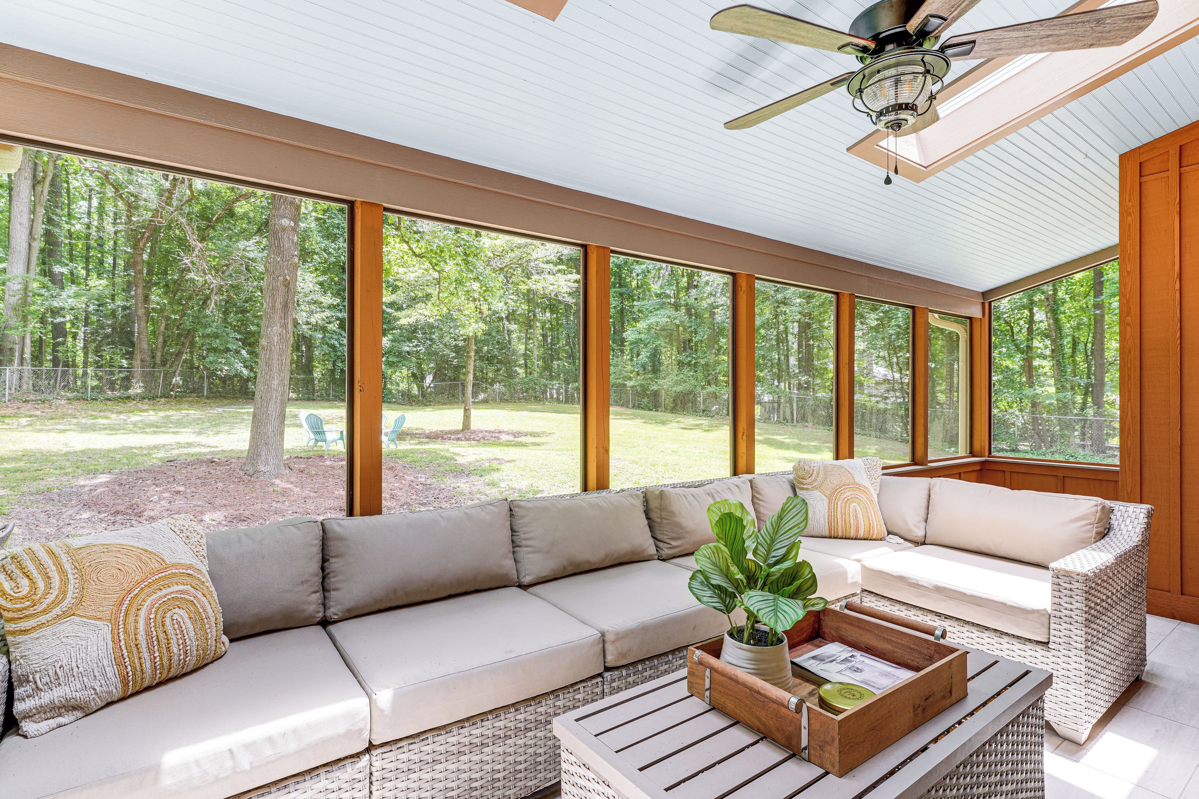 A sunlit room with a large window, a ceiling fan, and a wicker sofa with cushions.